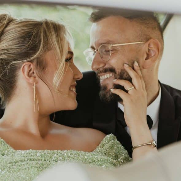 A couple smiles and looks at each other while sitting in a car's back seat.