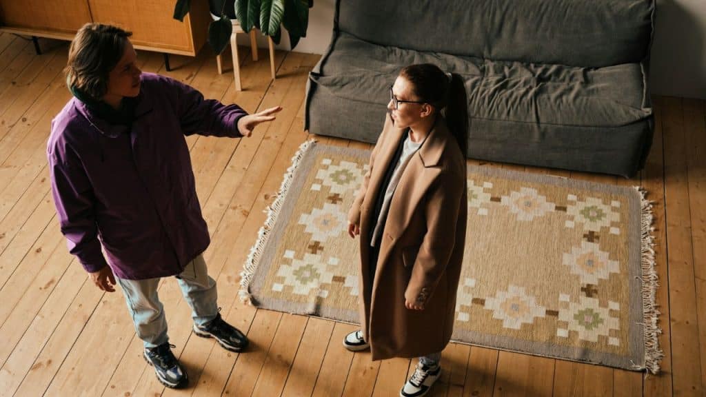 A couple in coats are standing on a rug in a wooden-floored living room, seemingly in a disagreement.
