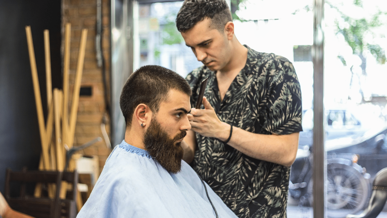 A barber in a floral shirt trimming a bearded man's hair with scissors and comb.