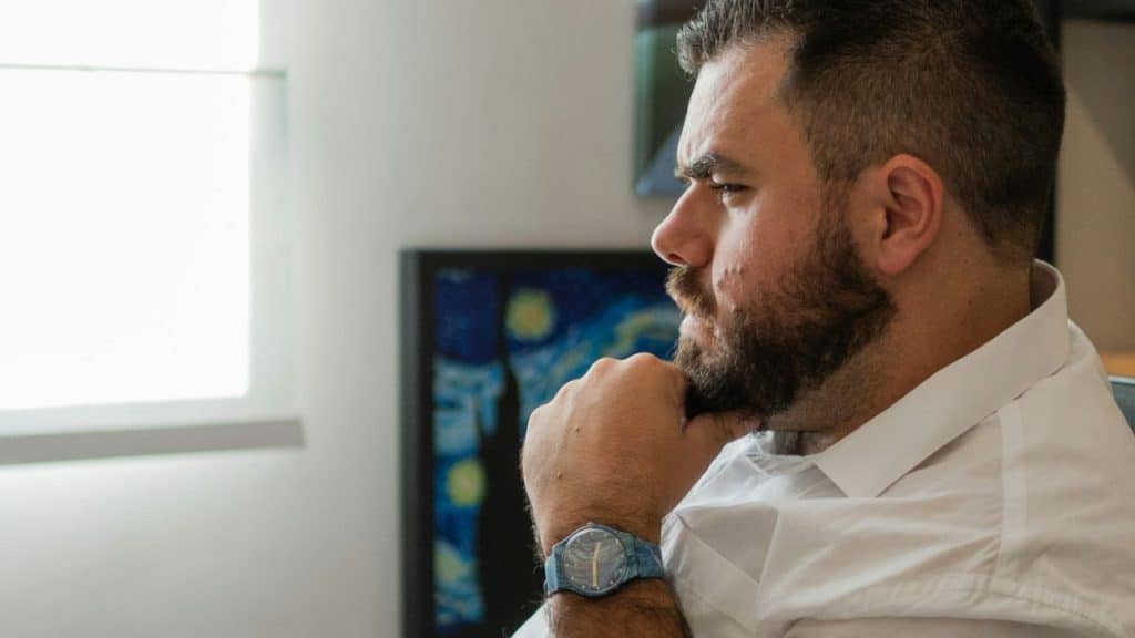 Bearded man in a white shirt and watch sitting thoughtfully, chin resting on his hands.