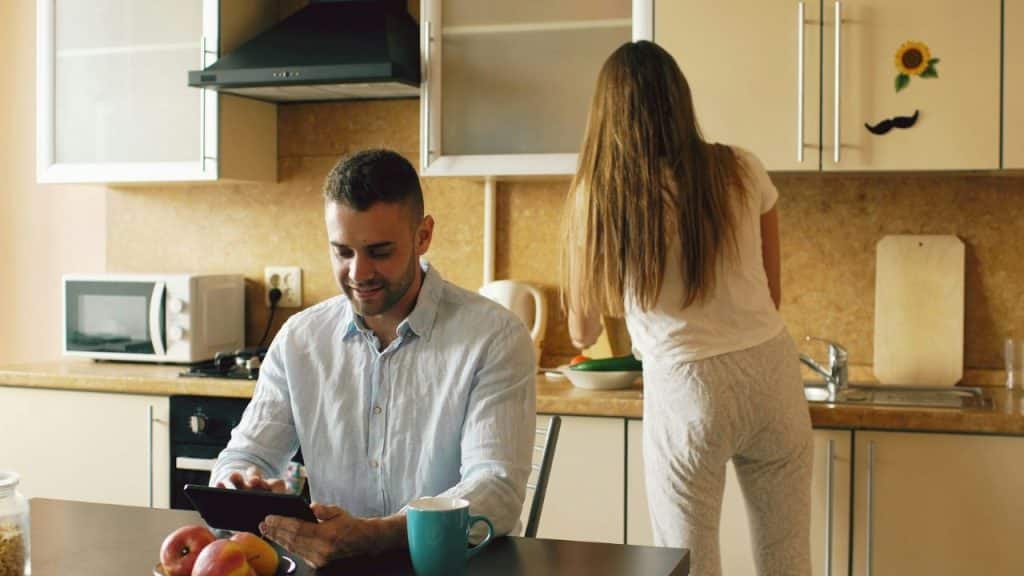 Man smiling at a tablet in a kitchen while a woman cooks with her back turned.