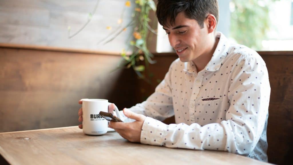 A smiling young man sits at a wooden table in a cafe, holding a white coffee mug and looking at his smartphone.