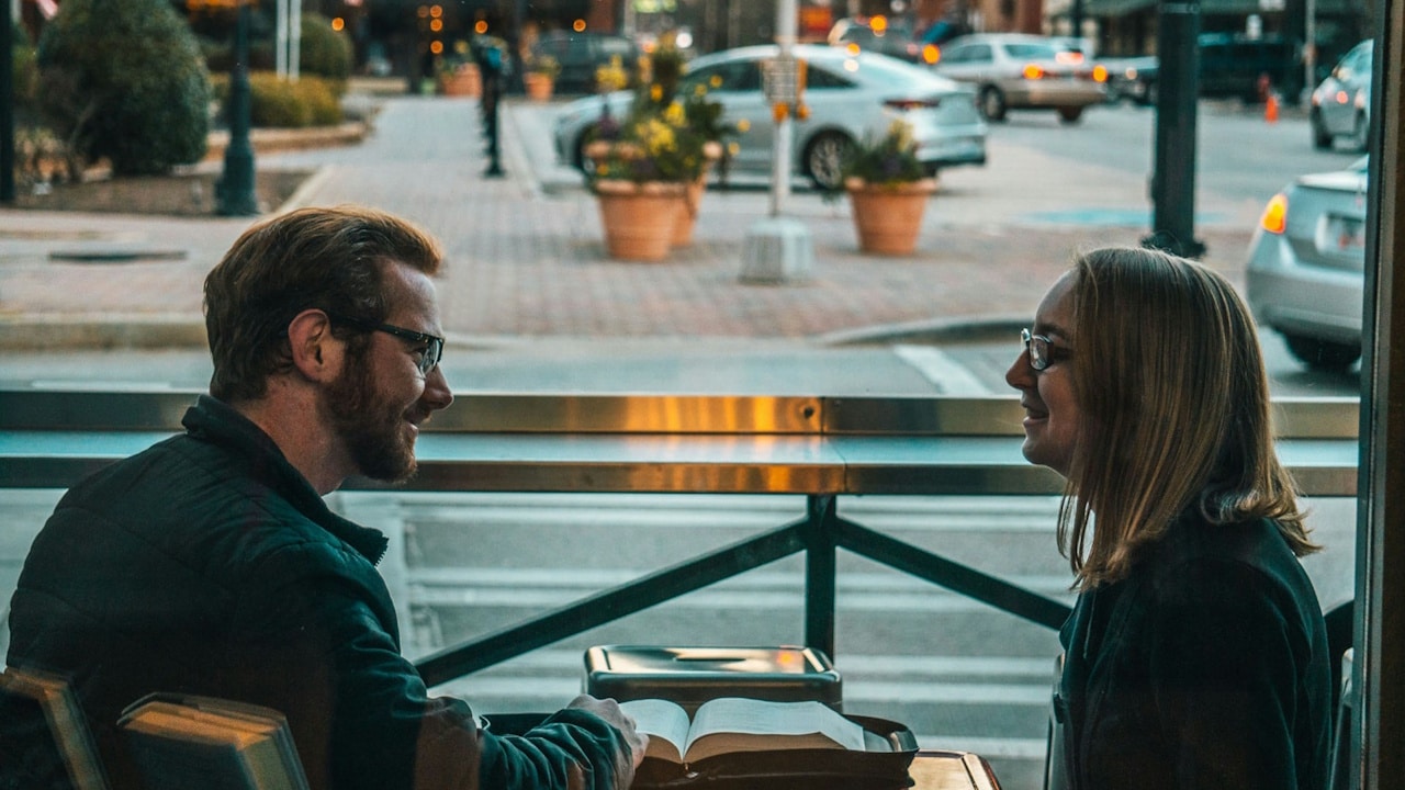 A smiling man and woman wearing glasses look at each other across a table with an open book.
