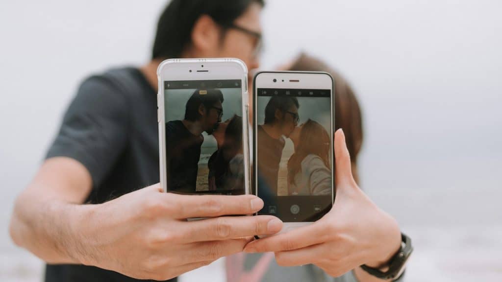 A close-up of a couple holding two smartphones showing a photo of themselves kissing, with the couple kissing in the blurred background.