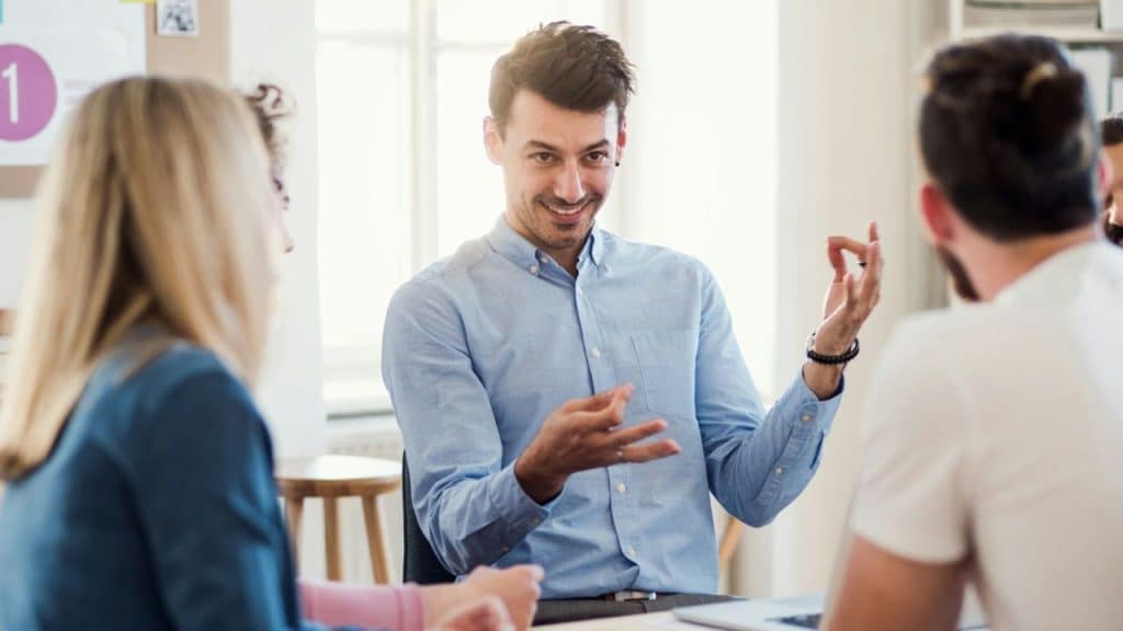 Man in a blue shirt gesturing and smiling while talking to two people in an office setting.