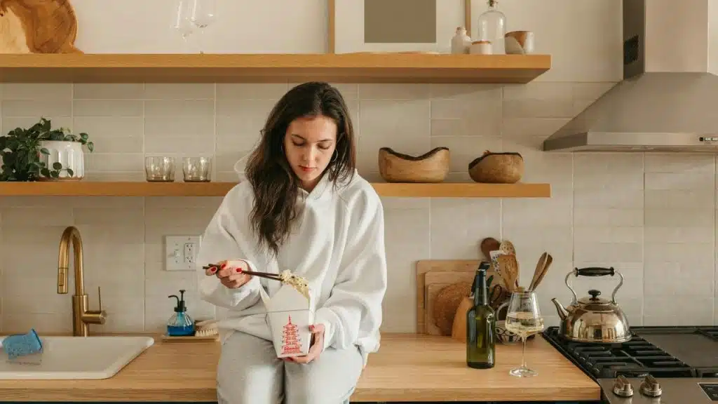 A woman sitting on a kitchen counter eating takeout noodles.