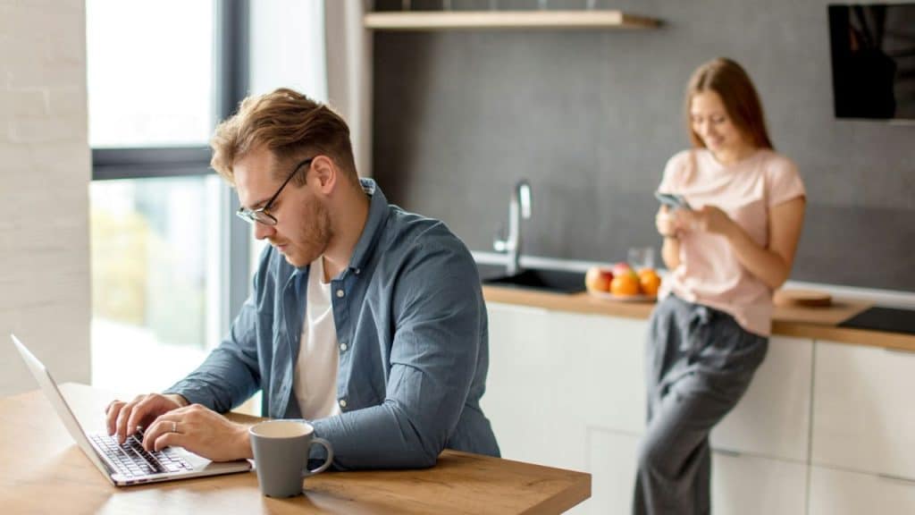 A man in a blue shirt and glasses is working on a laptop at a kitchen table while a woman uses her phone in the background.