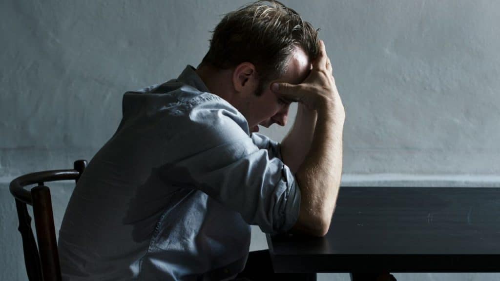 A distressed man sits alone at a dark table with his hands pressed to his head.
