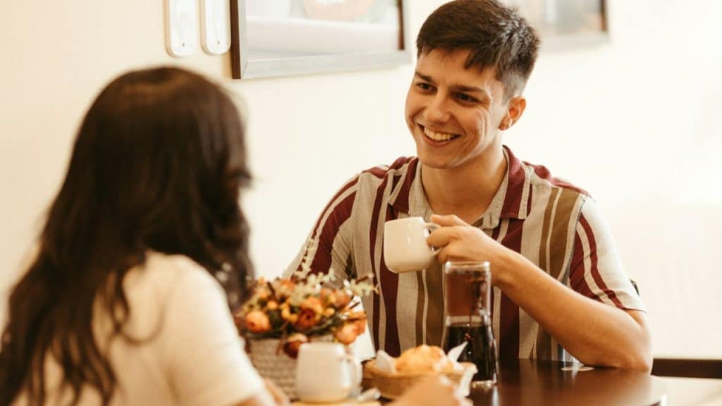Smiling man holding a coffee cup, talking to a woman across a table.