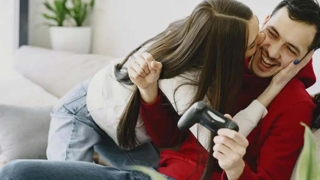 A woman playfully kissing a man on the cheek while he laughs and holds a game controller.