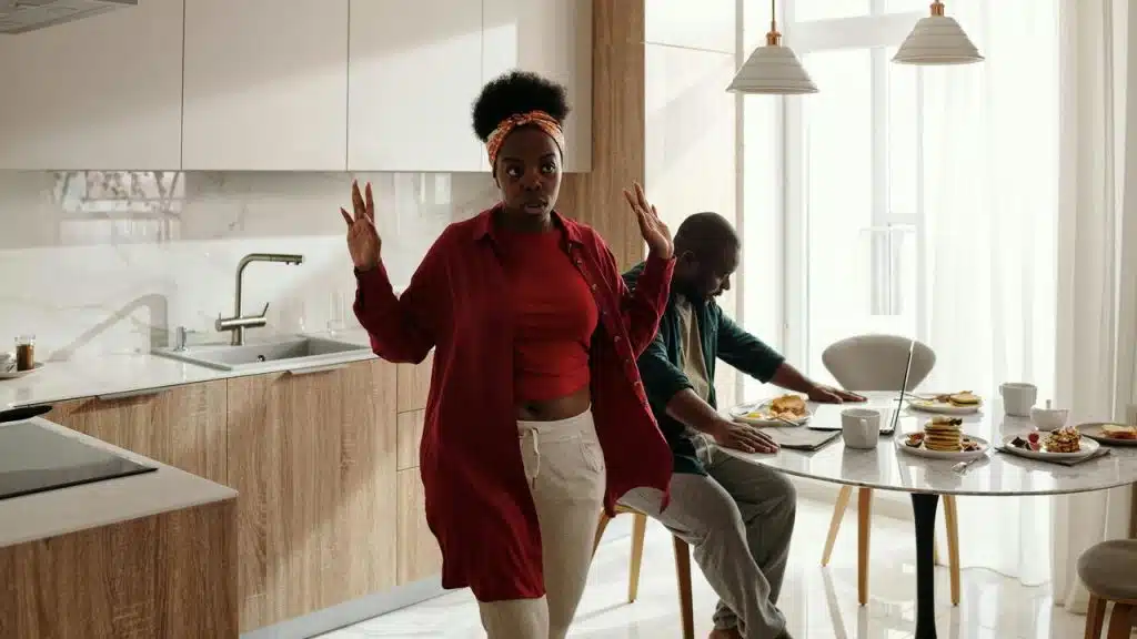 A woman raising her hands in a kitchen while a man eats at the table.