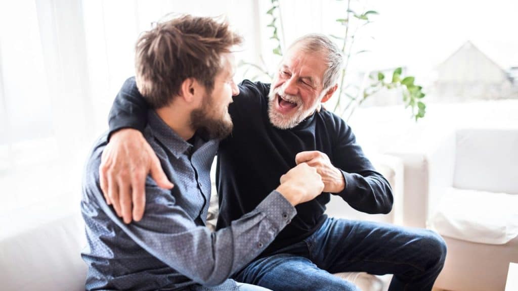 Happy elderly man hugging and laughing with a younger man indoors.