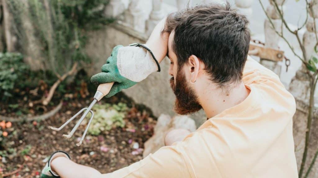 Man with a beard and earring holding a gardening tool, resting his head on his hand.