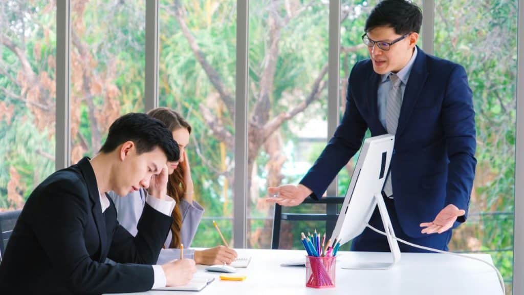 Man in a suit standing and gesturing to seated colleagues in an office.