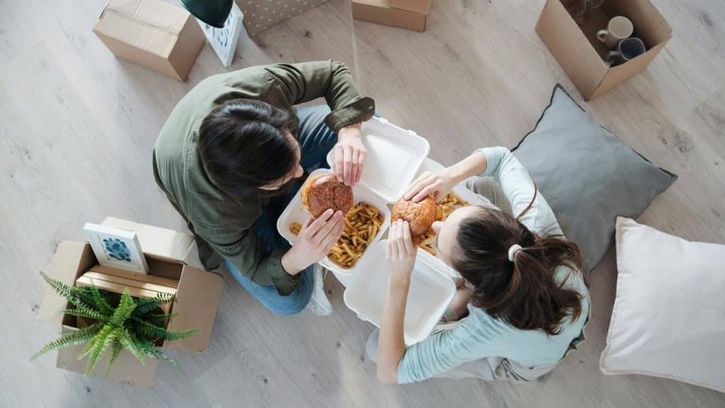 A couple eating burgers and fries on the floor among moving boxes.
