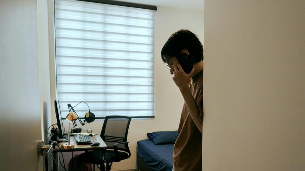 A man in a bedroom talking on a phone near a desk.