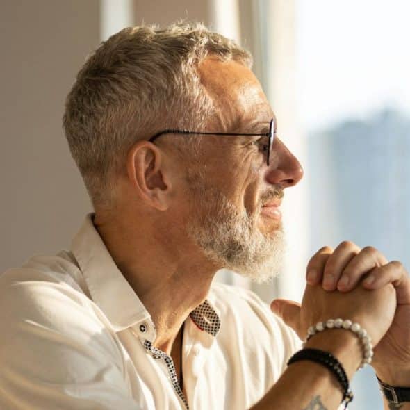 Gray-haired man in glasses and bracelets looking out a bright window.
