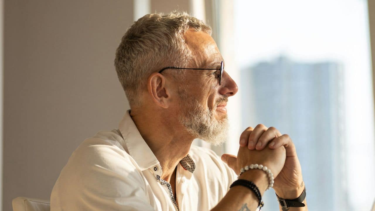 Gray-haired man in glasses and bracelets looking out a bright window.