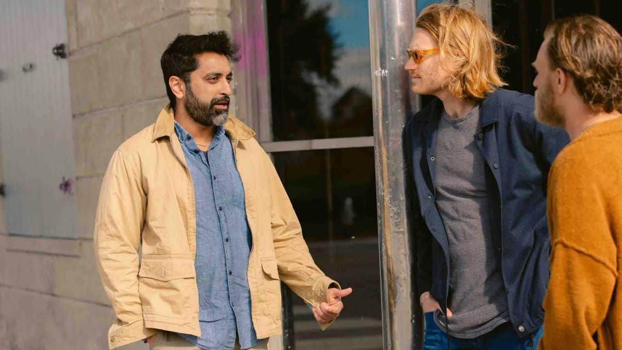 Three men stand outside talking; one wears a tan jacket and another has long blonde hair.