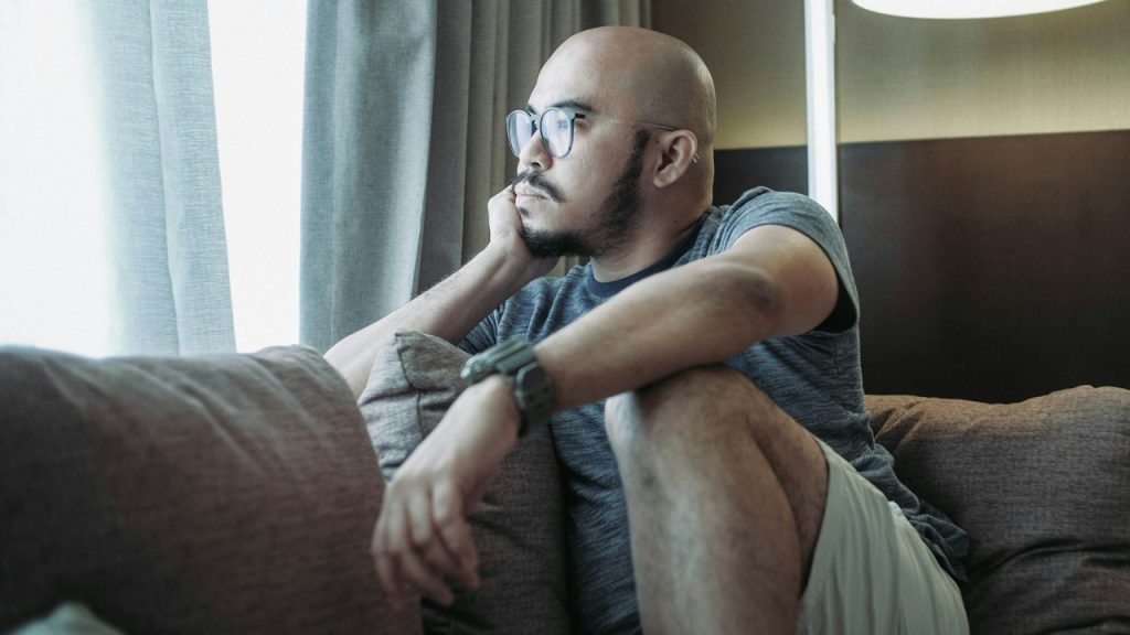 A bald man wearing glasses and sitting on a couch while looking outside a window.