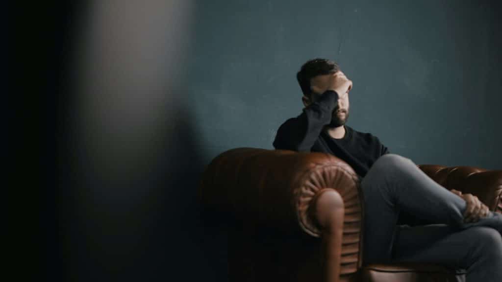 A man looking tired and holding his hand up to his face while sitting on a couch.