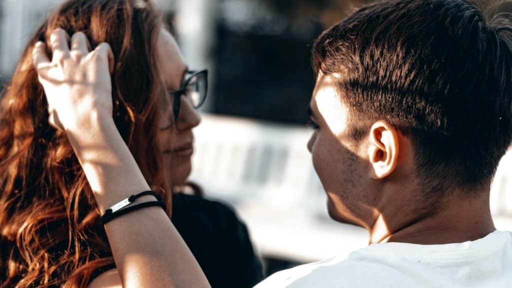 A close-up of a woman with auburn hair and glasses touching her hair while looking at a man in a white shirt.
