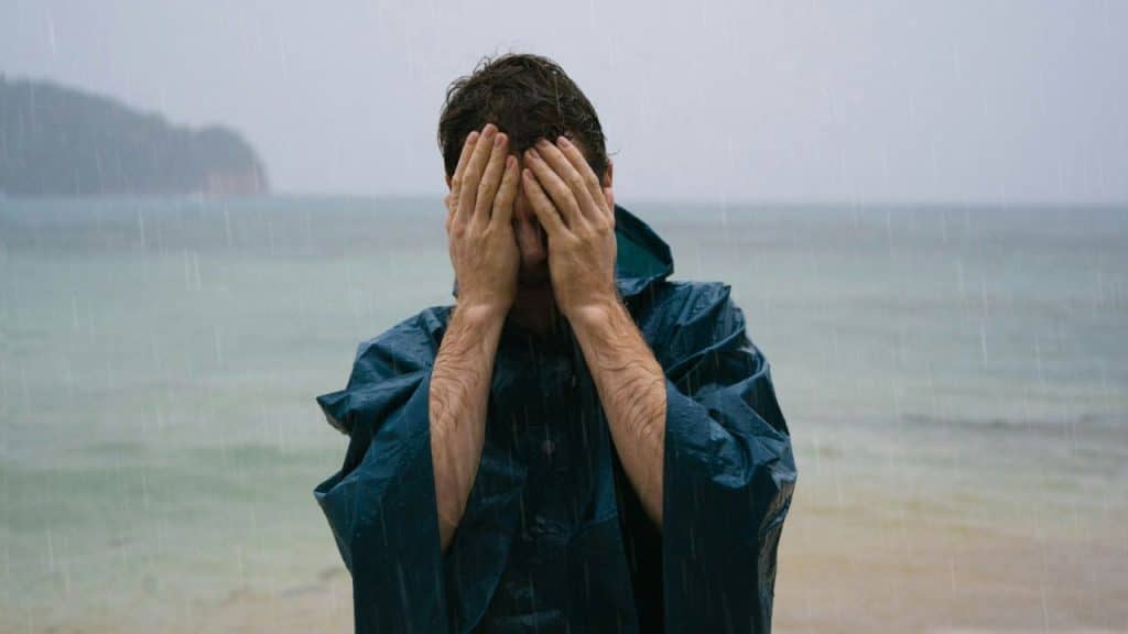 Man in a blue raincoat covering his face with his hands while standing in the rain by the ocean.