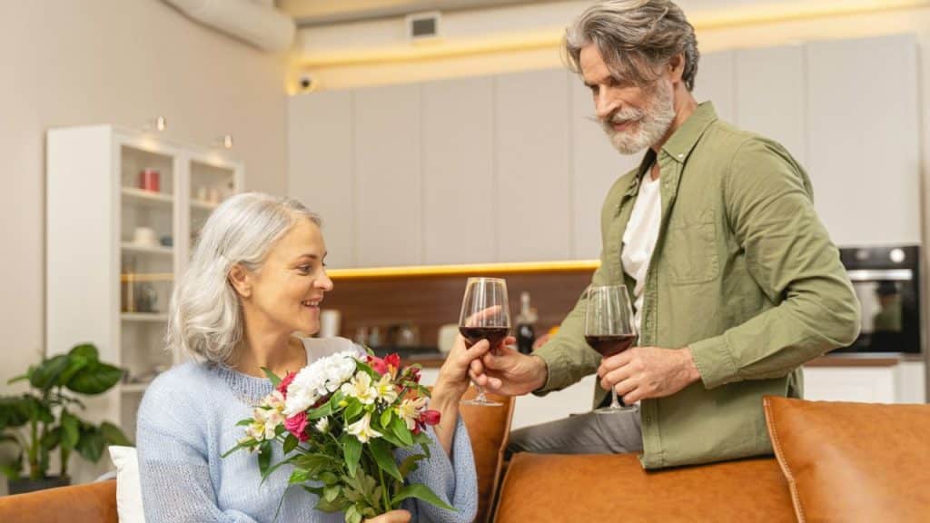 An older man and woman toast with wine glasses; she holds a bouquet of flowers.