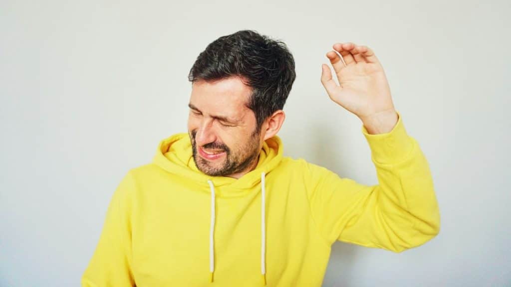 Man in a yellow hoodie grimacing and holding one hand up, against a plain background.