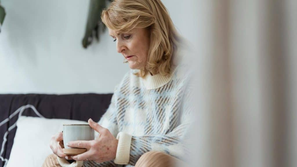 An older woman sitting on a couch holding a mug with a sad look.
