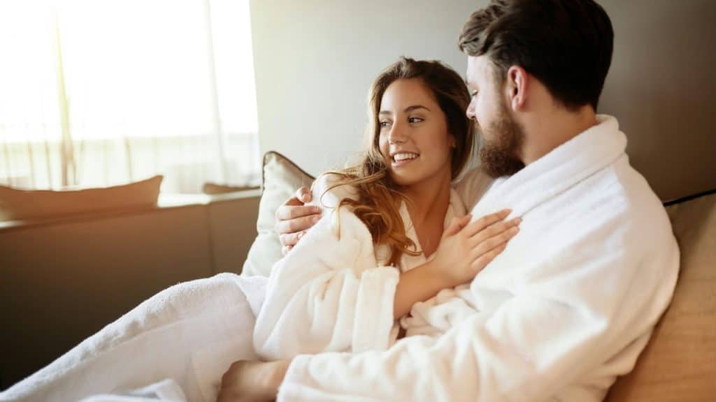 A smiling couple in white bathrobes embracing and looking at each other indoors.