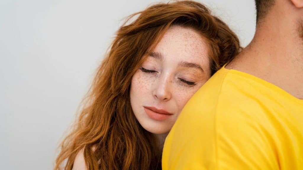 Close-up of a red-haired woman with freckles leaning her head on a man's yellow shirt.