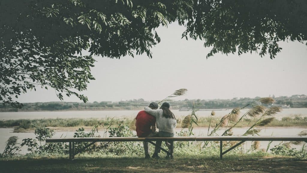 Couple sitting on a bench with their arms around each other, looking at a river.