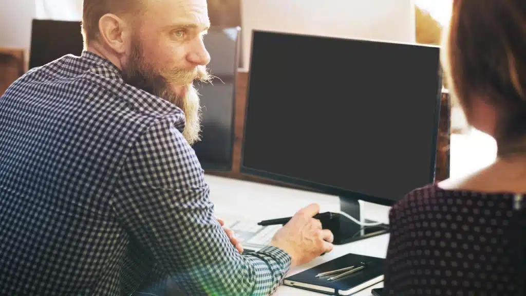 Bearded man pointing at a blank computer screen while talking to a colleague.