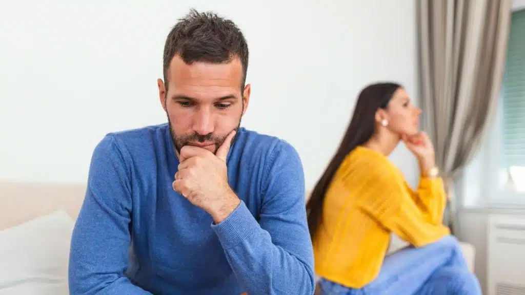 Man in blue sitting thoughtfully in the foreground while a woman sits away in the background.