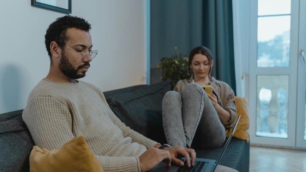 A man working on a laptop while a woman sits nearby using her phone.