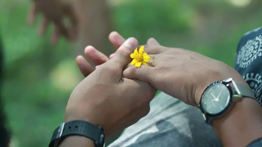 Close-up of two people holding hands with a small yellow flower resting on top.