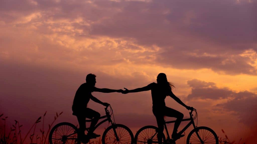 Silhouette of a couple holding hands while riding bicycles at sunset.