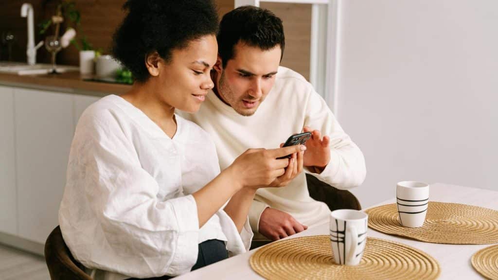 A couple sitting at a table, smiling while looking at a phone together.