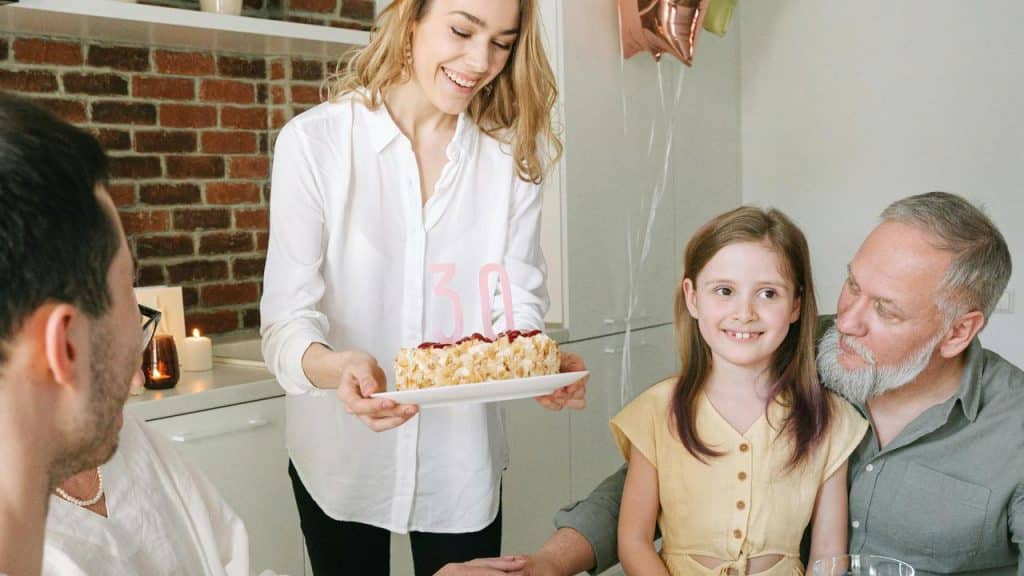 A smiling woman presents a birthday cake with the number 30 to a family at a table.