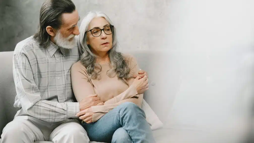 An elderly couple sitting on a couch, with the man comforting the woman who looks upset.