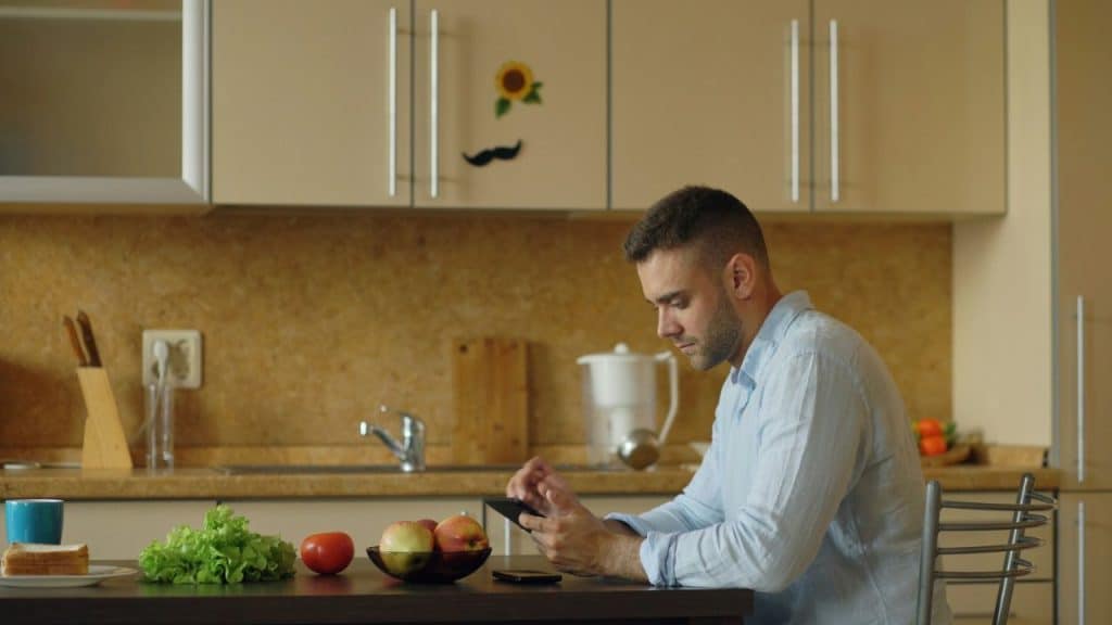 A man in a light blue shirt is sitting at a kitchen table, looking at his smartphone.