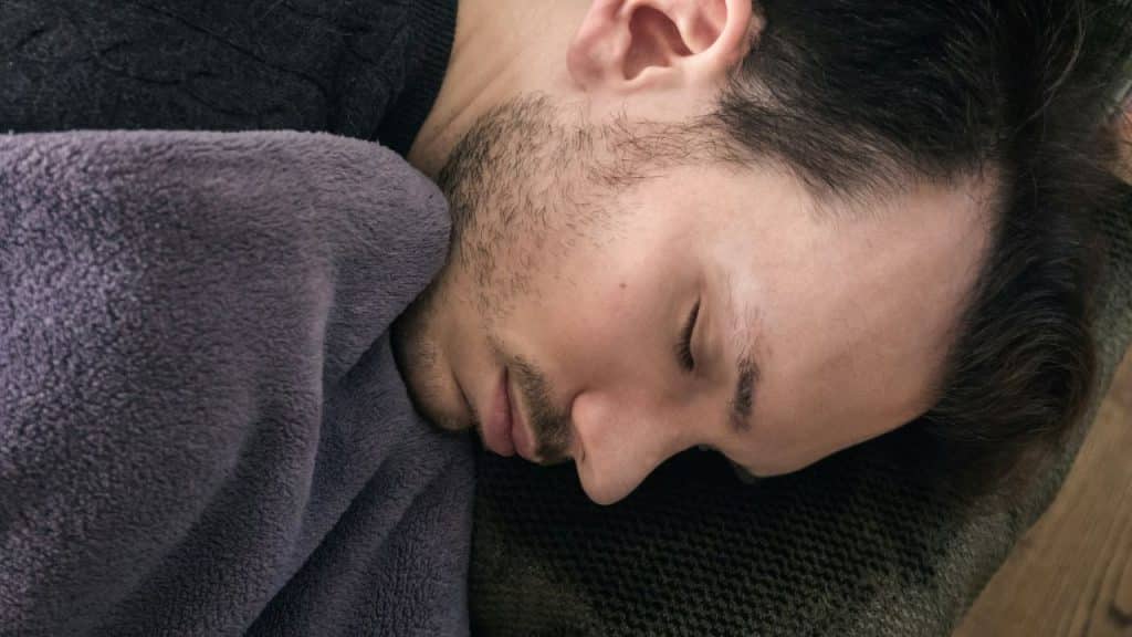 Close-up of a man with stubble sleeping under a purple blanket.