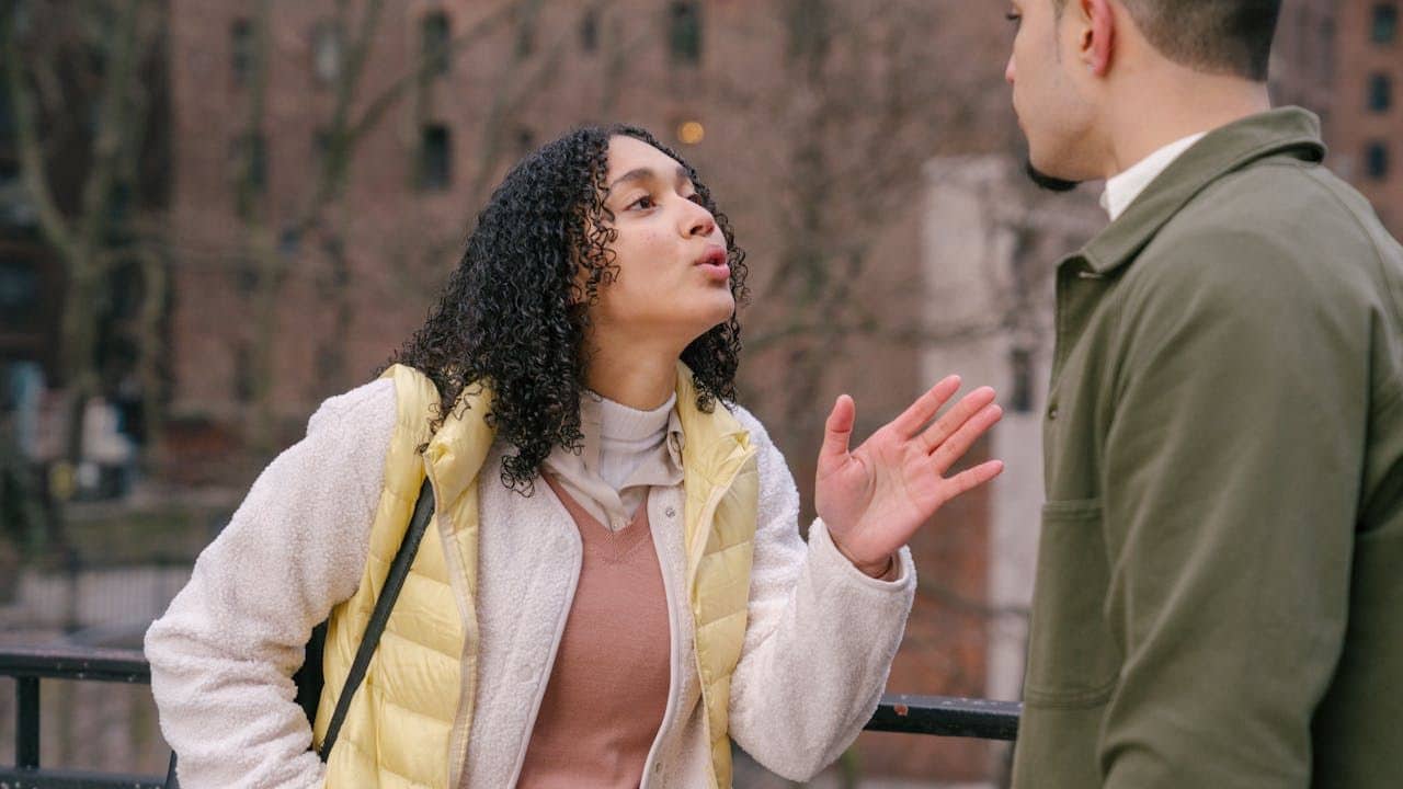 A curly-haired woman in a yellow vest is speaking animatedly and gesturing toward a man.