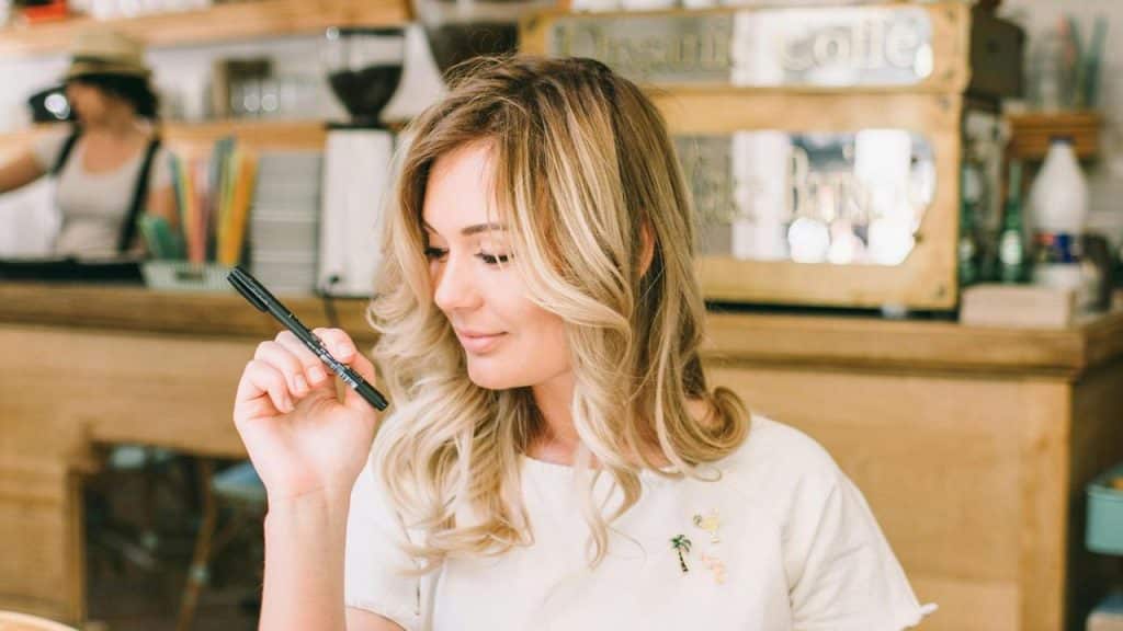 A woman sits in a café holding a pen and smiling thoughtfully.
