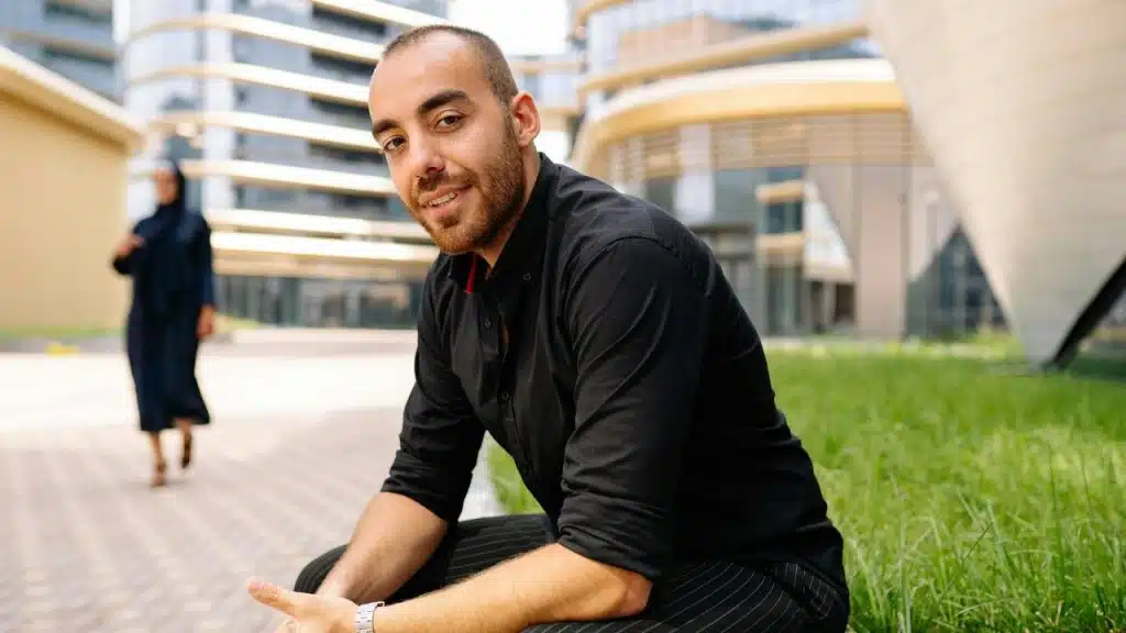 A man in a black shirt sitting outdoors in a modern urban area.
