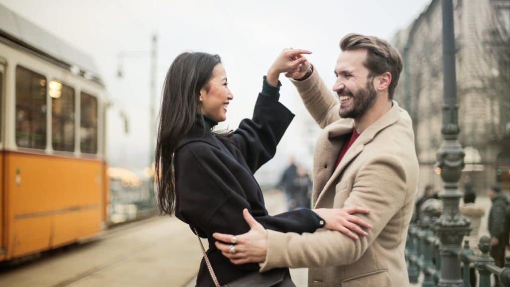 Happy couple dancing and laughing outdoors on a city street with a tram in the background.
