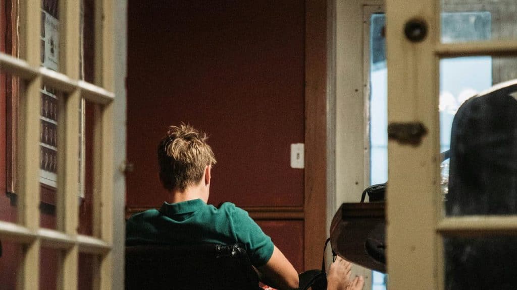 A person sits in a dimly lit room facing away from the camera near a desk.