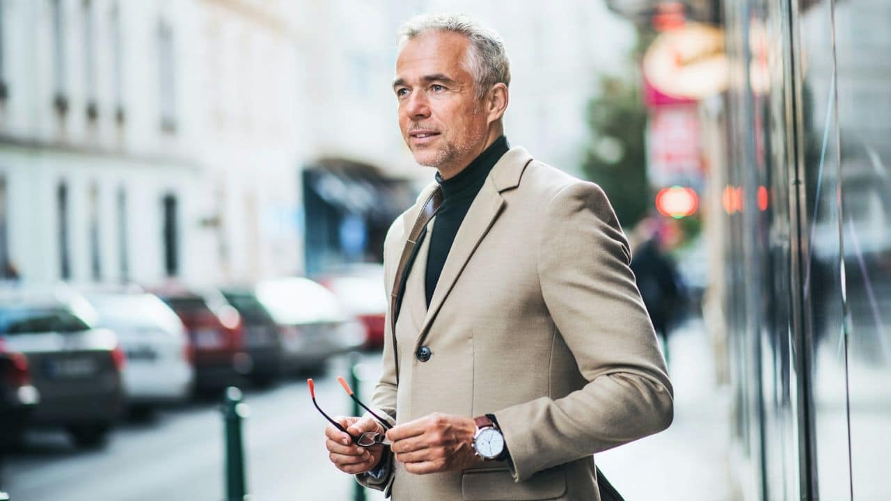 A distinguished, gray-haired man in a tan blazer is standing on a city street.