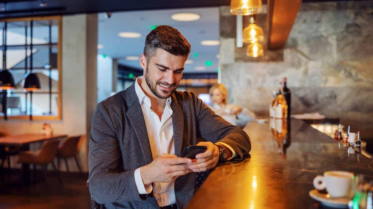 A smiling man in a suit jacket leans on a bar, looking at his smartphone.